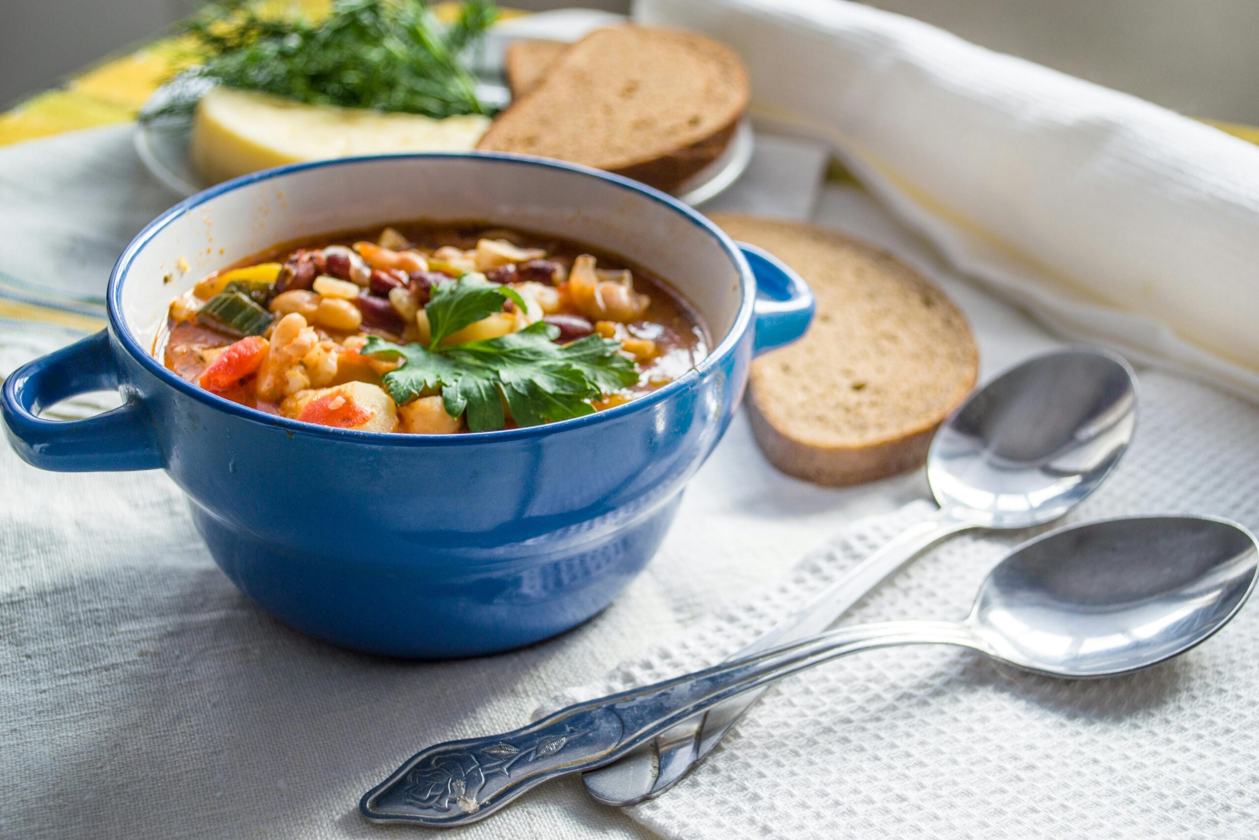 minestrone soup in a blue bowl with two spoons and sliced bread in the background